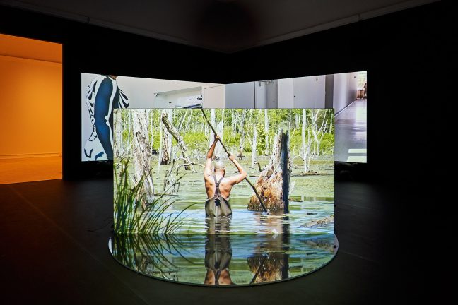 <p>A.K. Burns (U.S., b. 1975). <em>What is Perverse is Liquid (NS 0000)</em>. 2023. 3-channel HD video installation (color, 5.1 sound), 35:54 mins. Two projection screens, free-standing projection wall, plexiglass, sandbags, sand, rubber pool liner, and pennies. Courtesy of the artist. [Installation view, <em>A.K. Burns: What is Perverse is Liquid,</em> Henry Art Gallery, University of Washington, Seattle. 2024]. Photo: Jonathan Vanderweit.</p>
